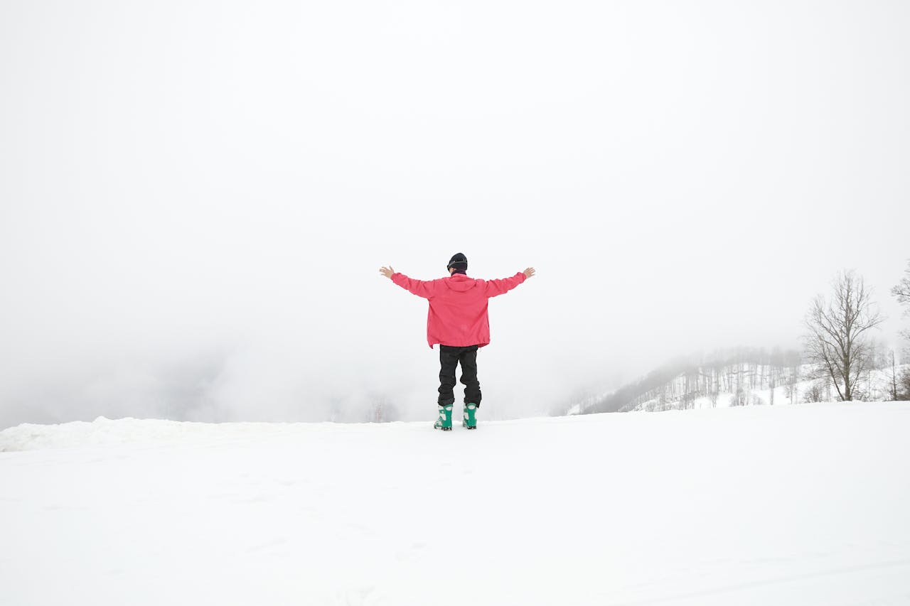 Osoba u roze jakni sa raširenim rukama stoji na snegu u planini.