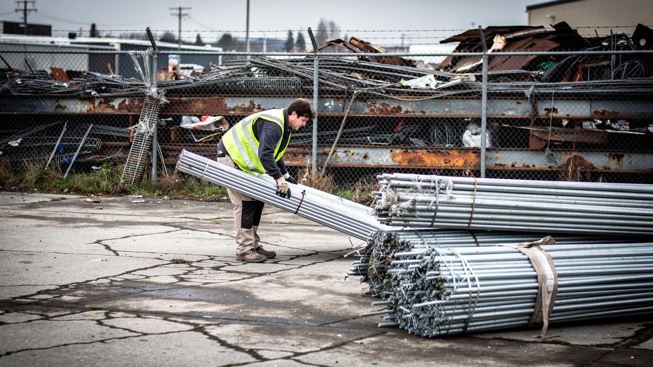 Radnik u fluorescentnom prsluku podiže snop metalnih cevi u industrijskom dvorištu