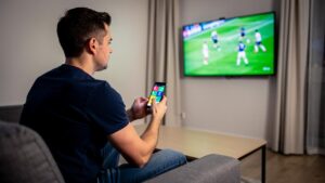 Muškarac sedi na kauču i koristi mobilni dok na televizoru ide fudbalska utakmica