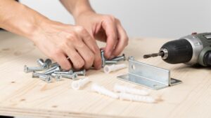 Ruke slažu metalne šrafove i plastične tiplove pored bušilice i metalnog ležišta